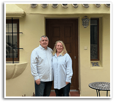 A man and woman standing in front of a yellow house. Description generated by AI