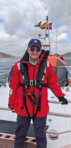 A man wearing a red jacket and a life vest is standing on a boat. Description generated by AI