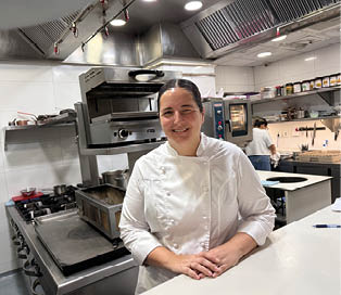 A woman in a white shirt and apron is smiling in a kitchen. Description generated by AI
