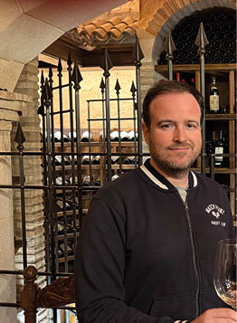 Two men standing in front of a wine cellar. Description generated by AI