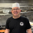 A man wearing a hat and apron is standing in a kitchen, smiling. AI generated content