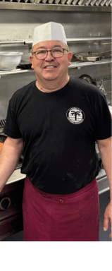 A man wearing a hat and apron is standing in a kitchen, smiling. AI generated content