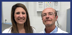Two adults wearing white coats stand side by side in a clinical setting with eye charts visible on the wall behind them. Description generated by AI