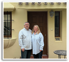 A man and woman standing in front of a yellow house. Description generated by AI