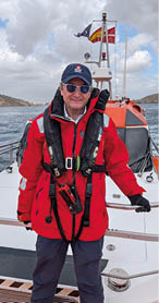 A man wearing a red jacket and a life vest is standing on a boat. Description generated by AI