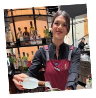 A woman in a black shirt and apron pouring a drink into a copper cup. Description generated by AI