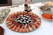A large platter of assorted sushi including salmon nigiri, tuna nigiri, and maki rolls is surrounded by bowls of shredded carrots, tuna salad, deviled eggs, grapes, and a cutting board with blue cheese on a white tablecloth. Description generated by AI
