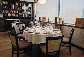 A round dining table set with plates, glasses, and a floral centerpiece is surrounded by five chairs in a warmly lit room with wooden floors and a display cabinet in the background. Description generated by AI