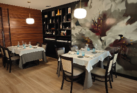 A modern dining area features two tables set with white and gray tablecloths, blue glasses, and neatly arranged plates and cutlery, with a large floral mural on one wall and wooden paneling on another. Description generated by AI