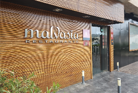A modern restaurant entrance with wooden paneling on the wall displaying the name “malvasia restaurante" next to glass doors and a tiled floor. Description generated by AI