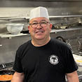 A man wearing a hat and apron is standing in a kitchen, smiling. AI generated content