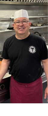 A man wearing a hat and apron is standing in a kitchen, smiling. AI generated content